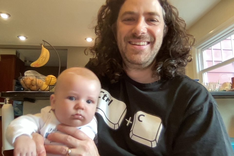 Max is wearing a black t-shirt with the keyboard keys CTRL + C shown. He has long brown curly hair. His youngest baby boy is in the photo with short fuzzy hair. 