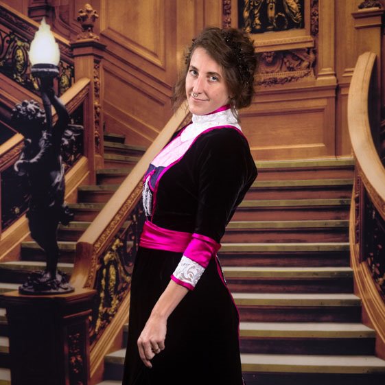 Stephanie wearing a black velvet gown with magenta trim and beading, standing in front of a backdrop of a staircase.