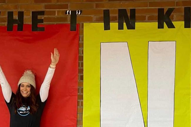 A student holds her arms above her head triumphantly in front of the word KIND, which is spelled out in bright bold primary colors in human sized poster paper. 