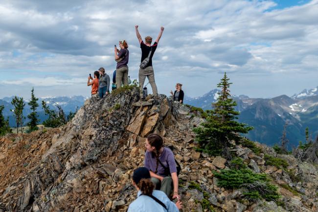 A student pumps their fists to the cloudy sky, as other students admire the Cascade mountain range from a craggy mountaintop.