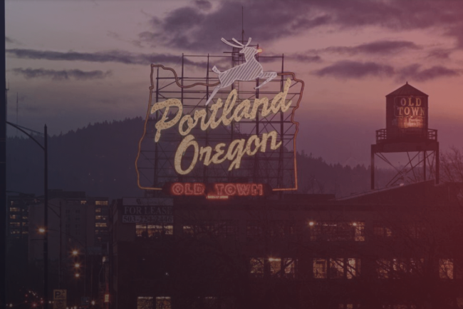 Portland city skyline at dusk. The Old Town sign with the state of Oregon outlined in neon and distinctive water tower are silhouetted against the evening skyline, glowing a dusky, dark-pink.