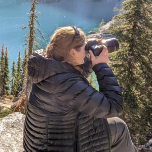 Jean in a puffy down coat sitting on a rock, looking over a turquoise lake below. She is holding a camera up to her eye taking a photo of the picturesque view below.  